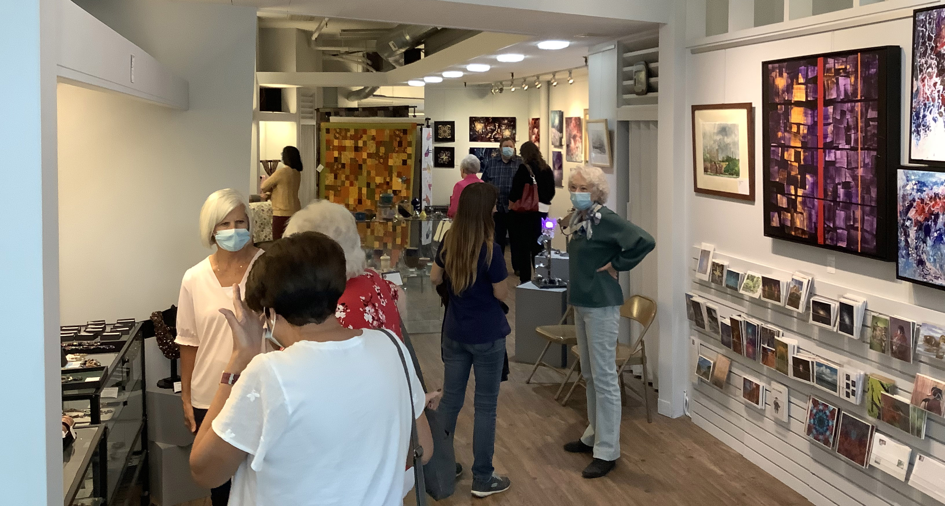 A crowd of masked gallery visitors, some conversing with featured artist Jan Crane.